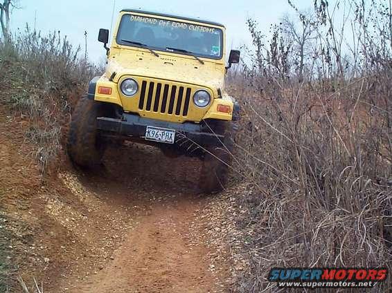 Yellow Jeep Wrangler on a dirt trail, front-left quarter view, with mud on tires
