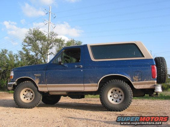 Blue Ford Bronco parked on dirt road, side view. Tan removable top and spare tire on back