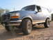 1996 Ford Bronco - A blue 1996 Ford Bronco with a tan stripe and lifted suspension sits on a dirt road.