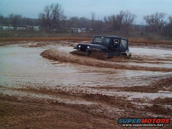 Black Jeep Wrangler driving through mud, front-left quarter view