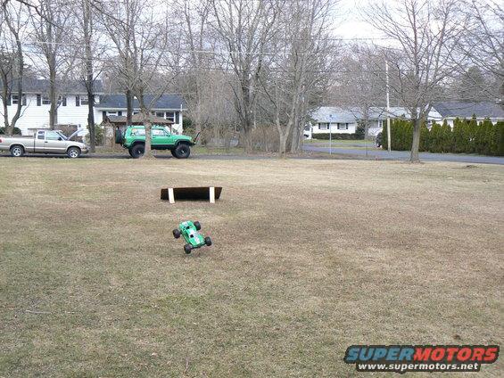 Green remote control car airborne over dry grass, with a silver pickup and a green SUV parked on the street behind it