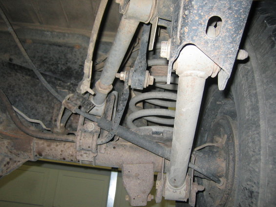 Rear suspension components of a vehicle, showing coil springs, shock absorbers, and control arms. The parts are dusty and show signs of rust