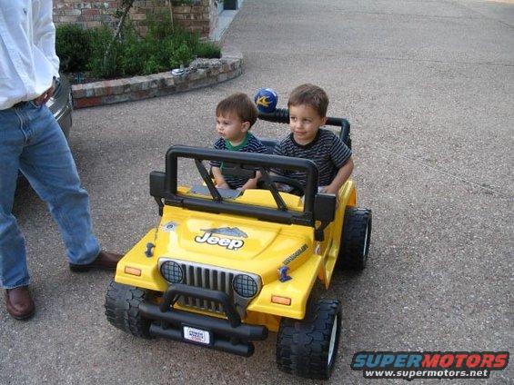Yellow toy Jeep with two young boys inside, parked on asphalt