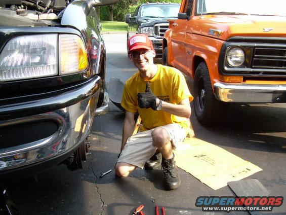 Man gives thumbs up next to a black truck and an orange truck. The orange truck appears to be a 1970s Ford