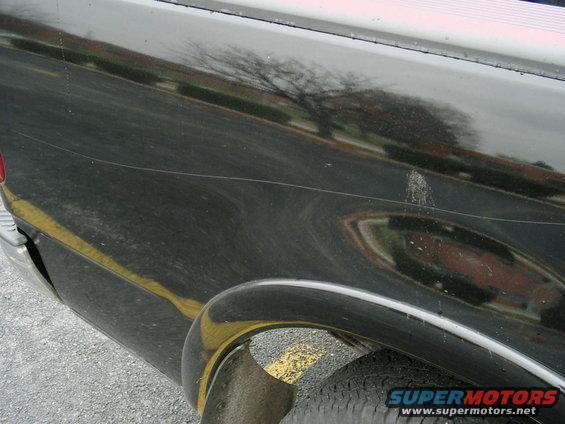 Black truck side panel with scratches and a yellow line, showing a reflection of a tree
