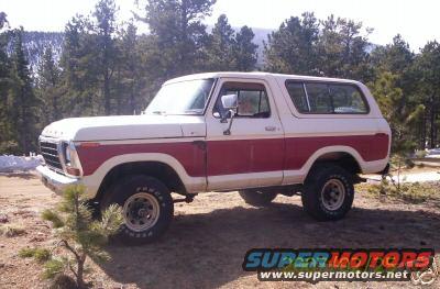 Two-tone red and white Ford Bronco parked on dirt with trees and mountains in background