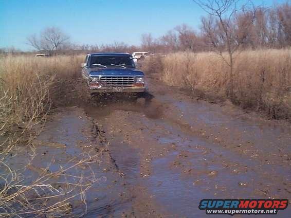 Blue 1978 Ford Bronco driving through deep mud on a dirt track, other vehicles visible in background