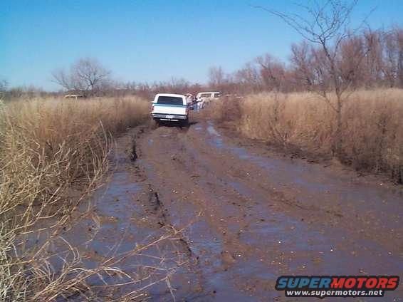 Several trucks driving down a very muddy, rutted road through dry grass and trees