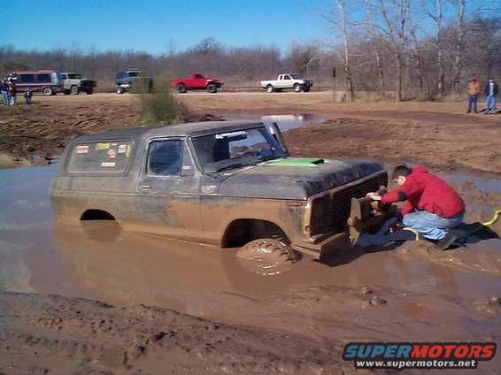 Mud-covered 1978 Ford Bronco partially submerged in thick mud, with a person in a red jacket attempting to winch it