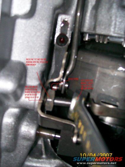 Close-up of a vehicle's transmission linkage with red text annotations pointing out a 7/8" nut and a detent spring