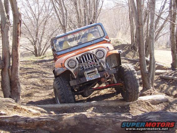 Orange Jeep Wrangler with large tires navigating an obstacle course of logs