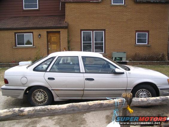Light-colored 1989 Ford Taurus sedan parked in front of a brick house, side view