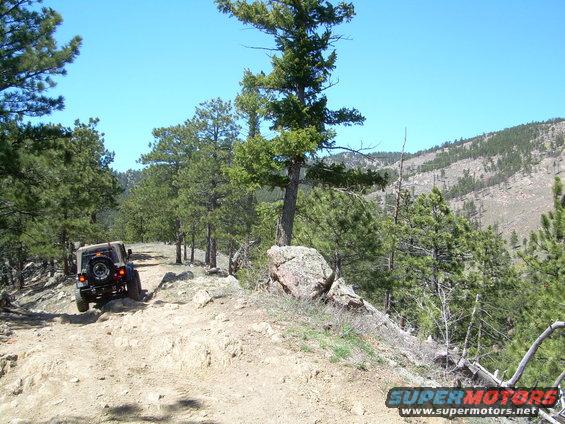 Jeep Wrangler driving up a rocky dirt trail with pine trees and a mountain in the background