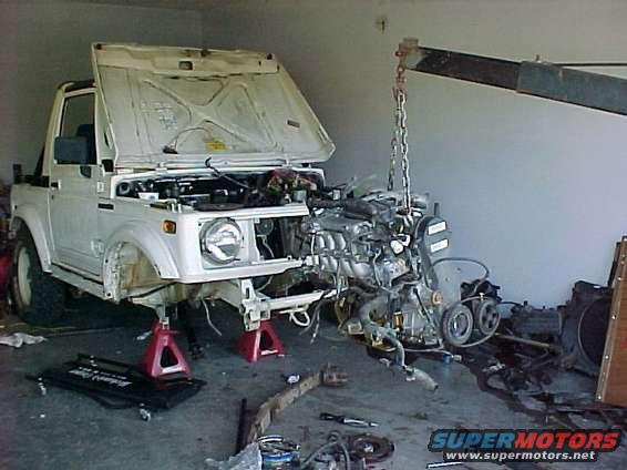 White Suzuki Samurai on jack stands, hood open, engine being lifted out by a hoist