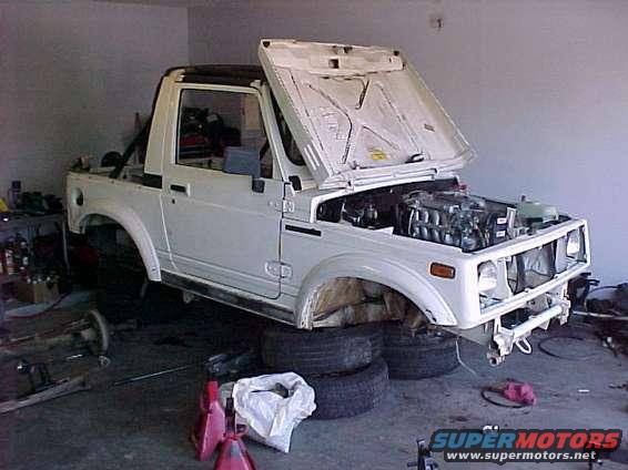 White Suzuki Samurai with hood open, engine exposed, on jack stands in a garage