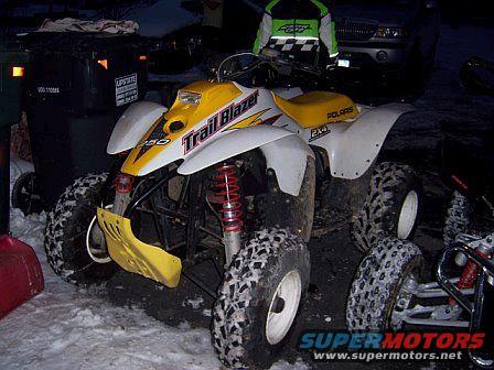 Yellow and white Polaris Trail Blazer ATV parked in snow, with a trash can and a car in the background