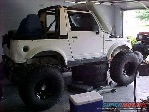 White Suzuki Samurai with large tires, parked in a garage. Another vehicle is partially visible to the right