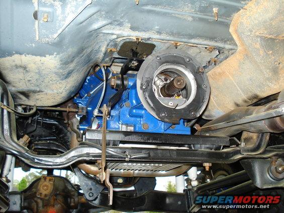 Underside view of a blue transmission with a transfer case output shaft visible. Rusty frame and suspension components