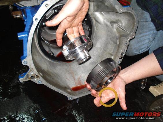 Hands holding transmission components, one shiny gear and a dark ring with a yellow seal