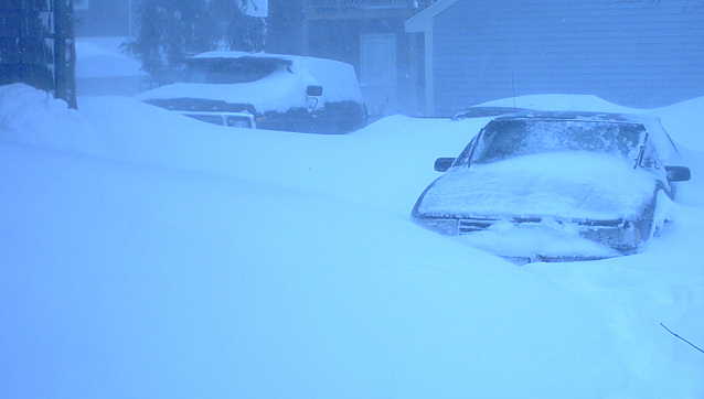 Two cars are almost completely buried under deep snowdrifts, with only the tops of their roofs and windshields visible