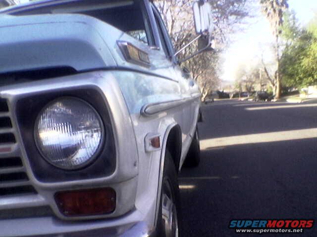 Close-up of a light blue and white pickup truck's front fender, headlight, and grille, parked on a street