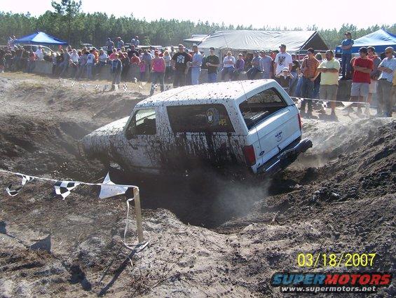 White SUV stuck in a mud pit, covered in mud, with spectators watching