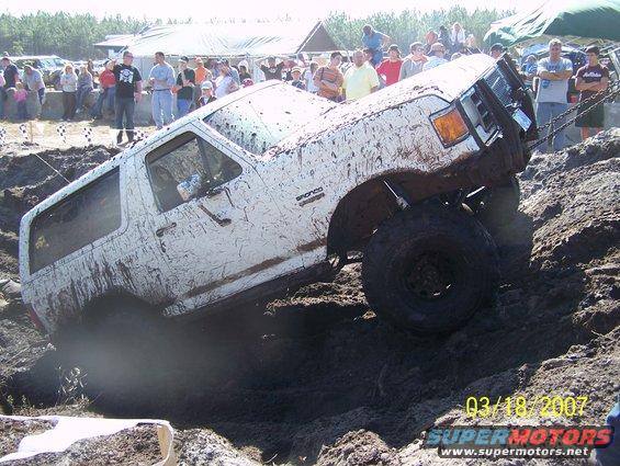 Mud-covered white Ford Bronco tilted on a dirt mound, front right wheel elevated