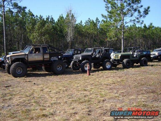 Modified pickup truck with large tires and custom cage, parked in a field with other off-road vehicles