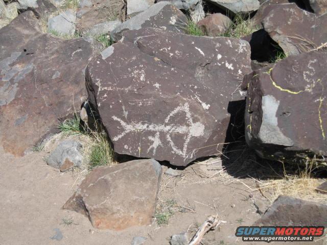 Petroglyphs of a bow and arrow on a dark rock surface, surrounded by other rocks and dry grass