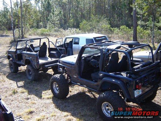 Several modified off-road vehicles parked in a wooded area. One is a dark-colored Jeep with a roll cage. Another is a dark-colored, older SUV body on a trailer chassis