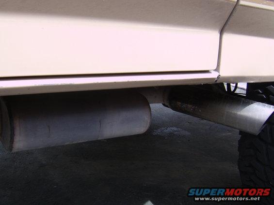 Cream-colored vehicle's lower body panel and dual exhaust pipes, one muffler visible