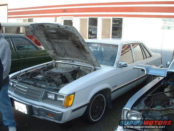 White 1980s sedan with hood open, showing engine. Another car is parked to the right