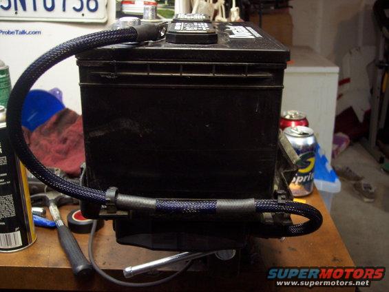 Car battery with braided cables attached, sitting on a workbench with tools and cans nearby