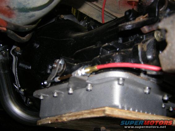 Close-up of a transmission pan with bolts and linkage. A red wire is visible