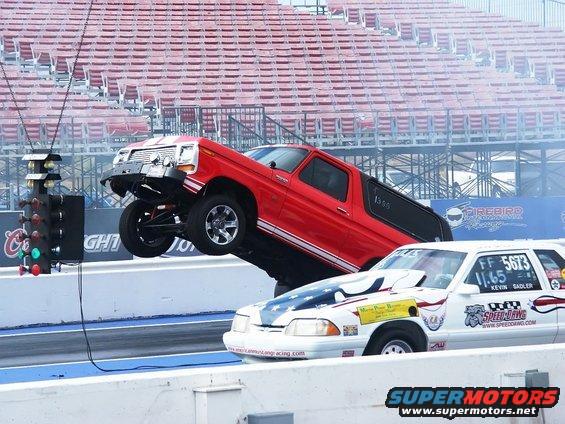 Red Ford Bronco performing a wheelie on a drag strip, with a white race car beside it
