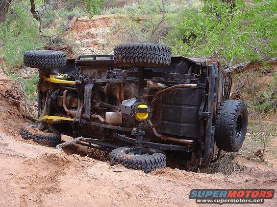 Upside-down off-road vehicle in sand and brush, showing undercarriage and suspension