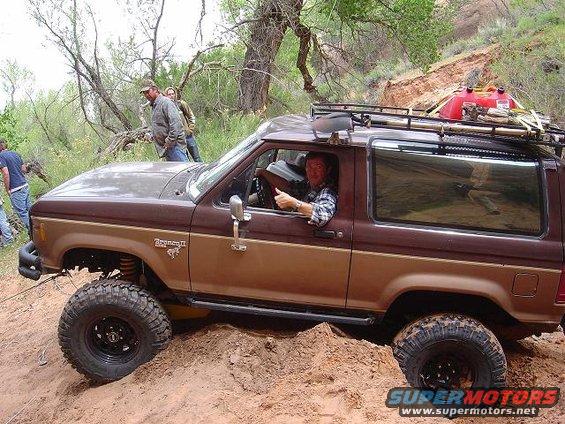 Brown Ford Bronco II with roof rack and extra gas can stuck in dirt, driver gives thumbs up