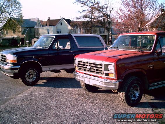 Two Ford Broncos parked side-by-side. The one on the left is dark blue with a grey top. The one on the right is red