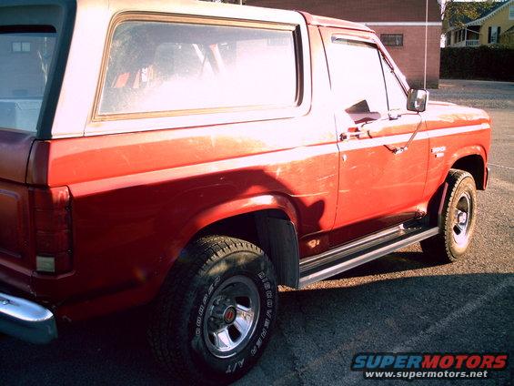 Red Ford Bronco with a white top, seen from the side at a 3/4 front angle. Chrome trim and running boards are visible