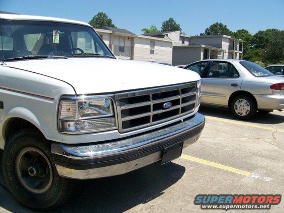 Front end of a white Ford F-Series truck with a chrome grille. A silver sedan is parked behind it