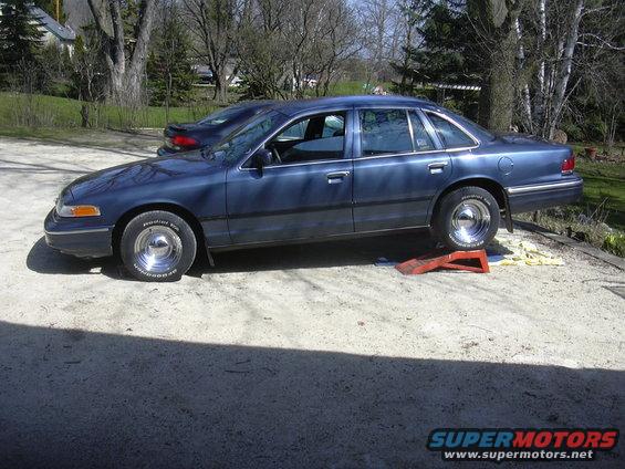 Blue sedan on ramps, one tire flat, chrome wheels, side view