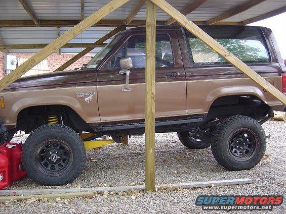 Brown two-tone Ford Bronco II lifted with off-road tires, parked under a wooden structure