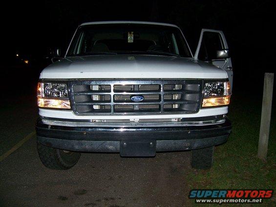 White Ford Bronco front end, driver's door open. Headlights on