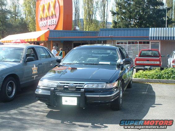 Black police car with push bar parked in front of a building with a XXX Root Beer sign. Another police car is to the left, and a red Chevrolet pickup is parked behind