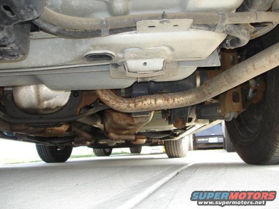 Underside view of a car's drivetrain and exhaust system. Another vehicle is visible in the background