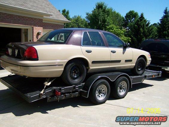Tan and brown 2003 Ford Crown Victoria on a dual-axle trailer, parked in front of a brick garage