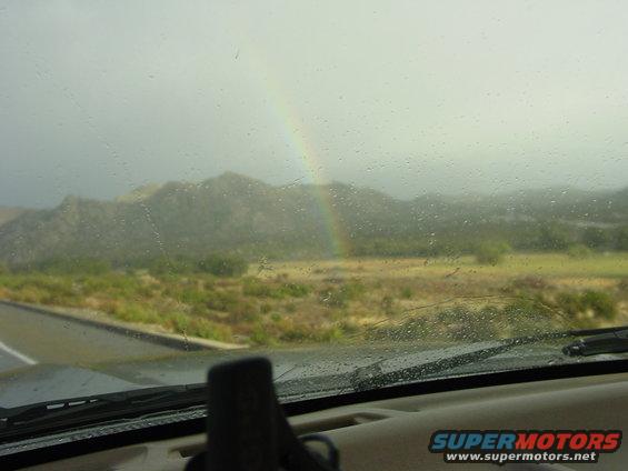 View of mountains and a rainbow through a windshield covered in raindrops. Supermotors.net logo visible