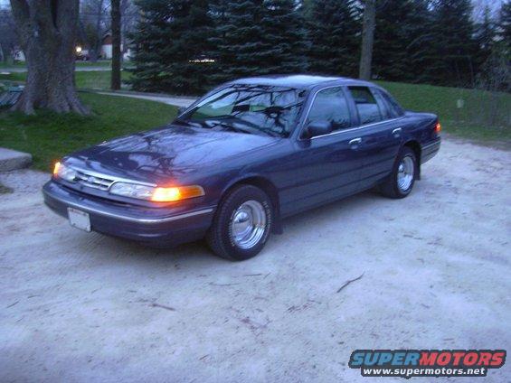 Dark blue sedan parked on a gravel driveway, front 3/4 view. Chrome wheels and amber turn signals are visible