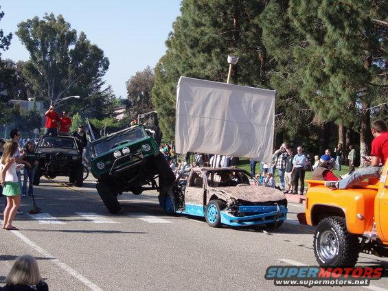 Green off-road vehicle on top of a damaged blue and brown car, with a large white sail structure