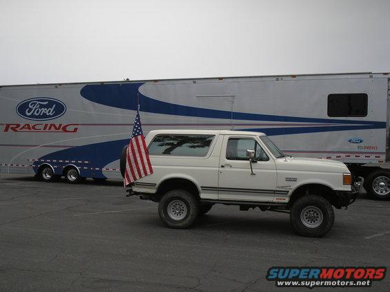 White 1990 Ford Bronco side view with American flag, parked next to a Ford Racing trailer